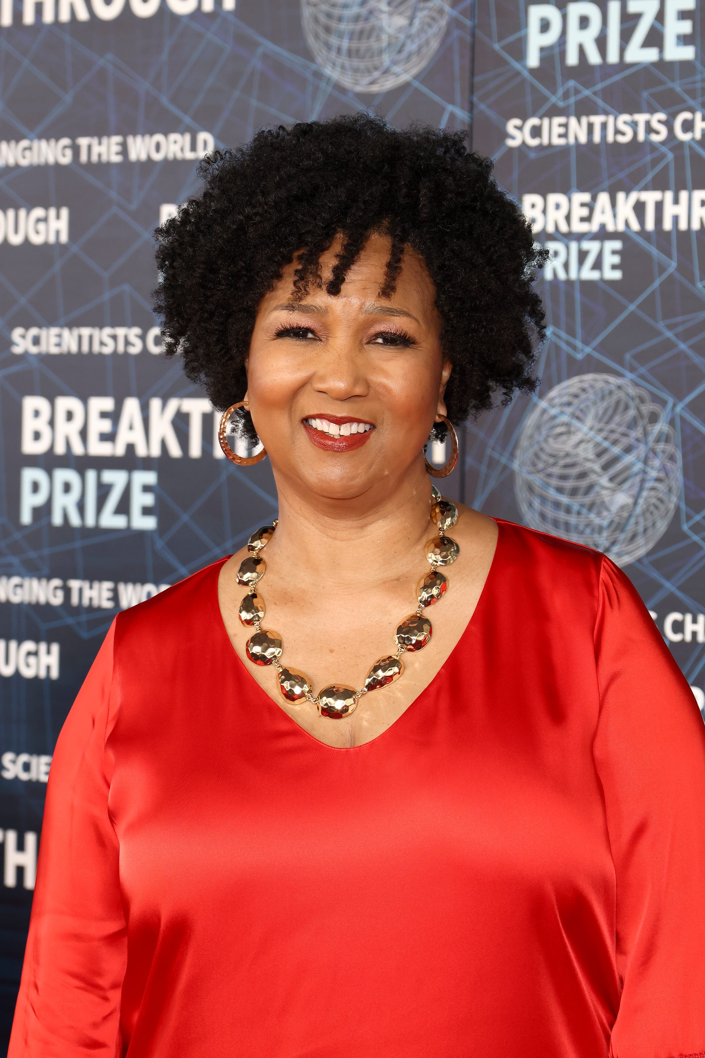 Mae Jemison in April 2023 mae jemison smiles at the camera while standing in front of a photo background with designs and writing, she wears a red top with gold hoop earrings a gold necklace